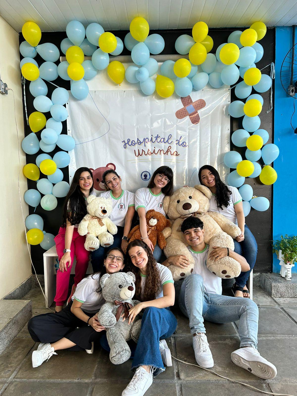 Integrantes da equipe sentados com ursinhos de pelúcia na entrada do evento, decorada com balões azuis e amarelos